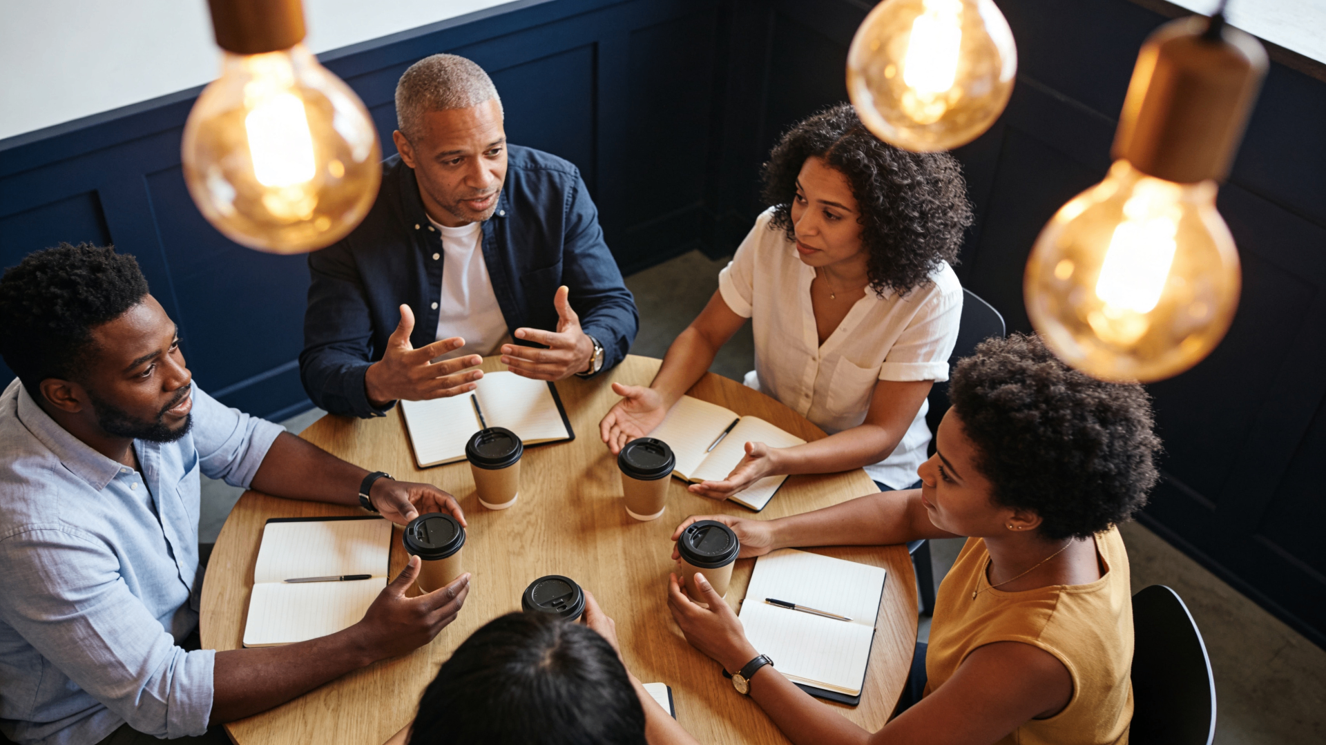 Group of professionals gathered around a wooden roundtable in conversation under warm Edison bulbs